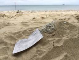A small square of paper with pencil rubbing on it sits on the sand with sea in distance.