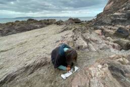 Image of Ruth Broadbent drawing on rocky beach with sea in background (2021)